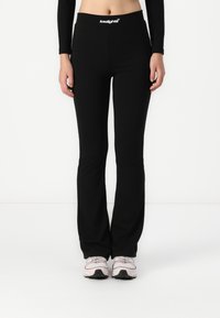 Leggings noires évasées avec une large ceinture comportant un logo. Le tissu est lisse et extensible, associé à des chaussures de sport légères.