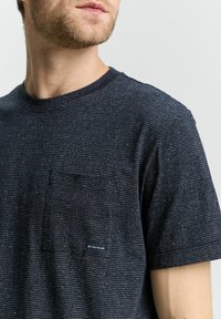 Camiseta de rayas azul oscuro con cuello redondo, mangas cortas y un pequeño bolsillo en el pecho. Presenta una tela texturizada y una etiqueta con el logotipo.