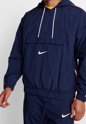 Marineblå vindjakke med hvidt Nike-logo, halv-zip front, kengurupocket, elastiske manchetter og røde sidelæbestykker. Fremstillet i letvægtsmateriale.
