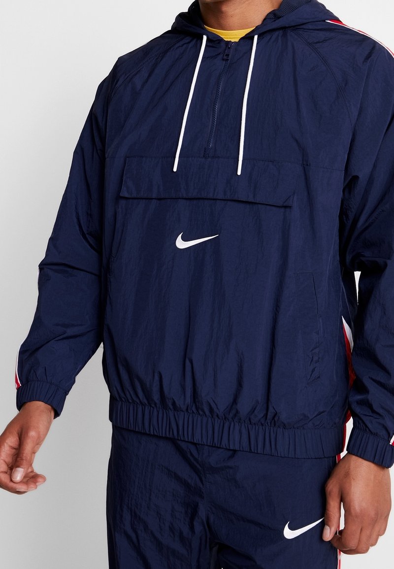 Marinblå vindjacka med vitt Nike-logotyp, halvdragkedja fram, känguruficka, elastiska ärmslut och röda sidopartier. Tillverkad av lättviktsmaterial.