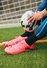 Botas de fútbol de color rosa brillante con material de malla y acentos negros, descansando sobre césped verde, al lado de un balón de fútbol multicolor.