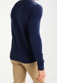 Man med en figursydd marinblå långärmad tröja och beigefärgade byxor som står lätt vänd bort från en enkel vit bakgrund.