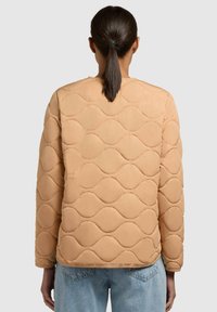 Giacca trapuntata beige con motivi a onda, caratterizzata da spalle arrotondate, maniche lunghe e orlo dritto, abbinata a jeans azzurri chiari.
