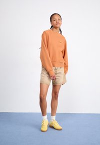 Orange sweatshirt med ribbede manchetter og kant, beige denim shorts og gule sneakers med hvide sokker. Model står på blåt gulv.