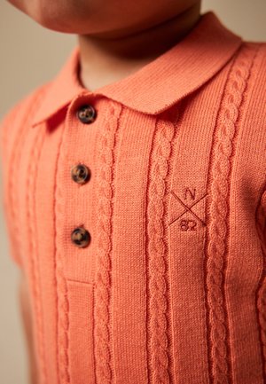 Zbliżenie dziecka ubranego w koralową dzianinową koszulkę polo z warkoczowymi wzorami, brązowymi guzikami i haftowanym "N 82" na piersi.