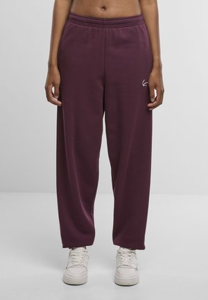 Bordó melegítőnadrág elasztikus derékpánttal, bő szárú szabással és finom logórészlettel. Puha anyag sima tapintással. Fehér tornacipő alatt.