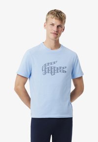 T-shirt en coton bleu clair avec un logo de crocodile géométrique en bleu foncé. Manches courtes, col rond et coupe standard.