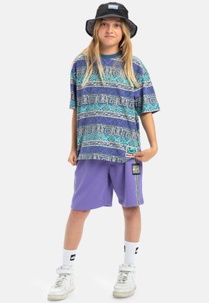 Paarse shorts met zijaccenten, oversized shirt in blauw en paars met patroon, witte sneakers en een zwarte bucket hat met logo.
