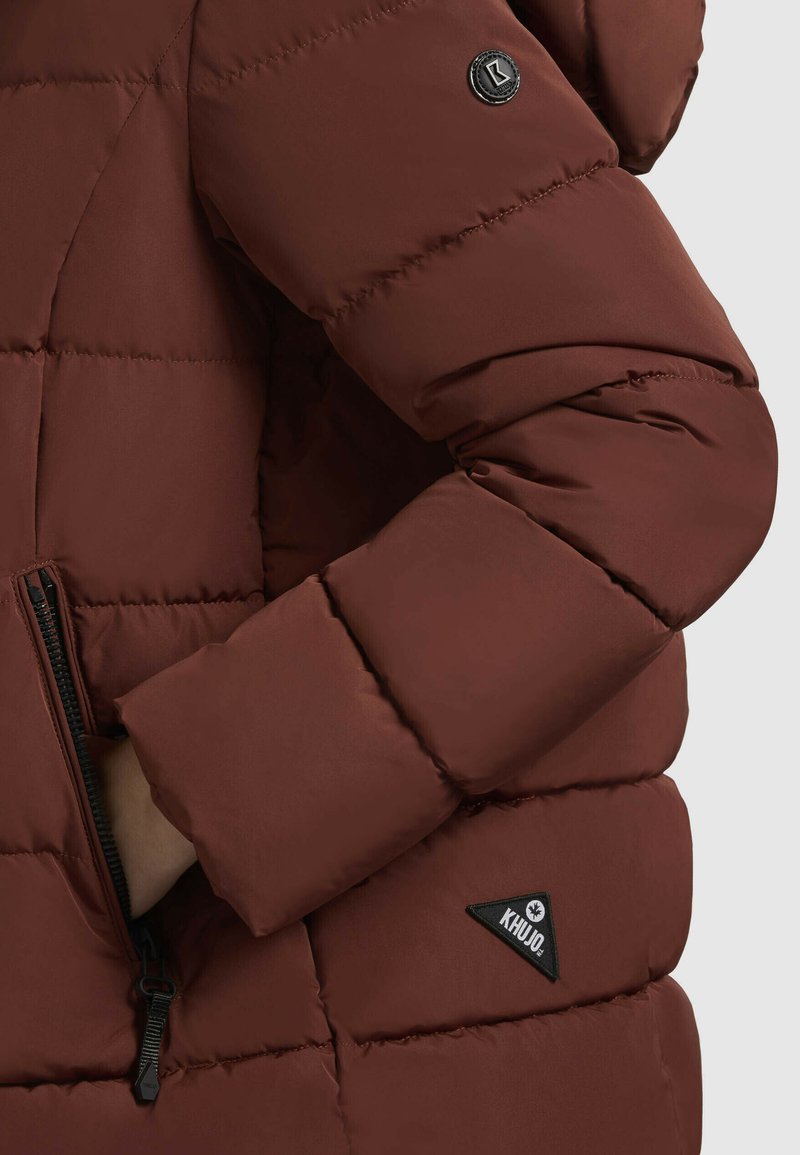 Veste rembourrée marron avec un design matelassé, dotée d'une poche zippée, d'un patch logo noir et d'une finition texturée sur le tissu.