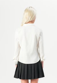 Bijela košulja dugih rukava s zaobljenim rubom i detaljima na manžetama, uparena s crnom plisiranom suknjom. Glatka tekstura, minimalistički dizajn.