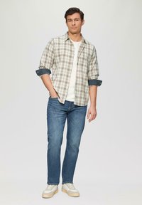 Beige und grün karierte Button-Up-Hemd, darunter ein weißes T-Shirt, blaue Jeans und weiße Sneakers mit beigen Akzenten.