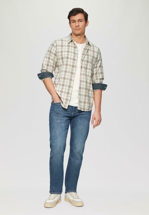 Licht geruite knopenoverhemd in beige en groen, witte T-shirt eronder, blauwe spijkerbroek en witte sneakers met beige accenten.