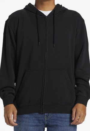 Persoon draagt een zwart vest met ritssluiting, voorzakken en koordjes, gecombineerd met een blauwe spijkerbroek, getoond van nek tot heupen.