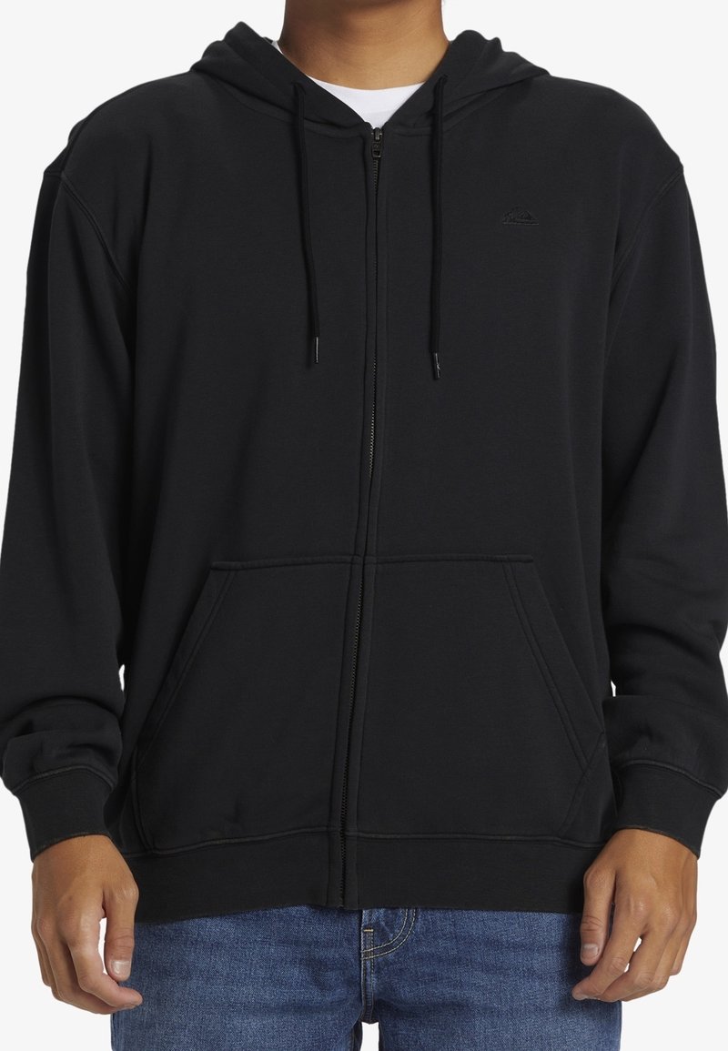 Personne portant un sweat à capuche noir zippé avec poches avant et cordons, associé à un jean bleu, visible du cou jusqu'aux hanches.