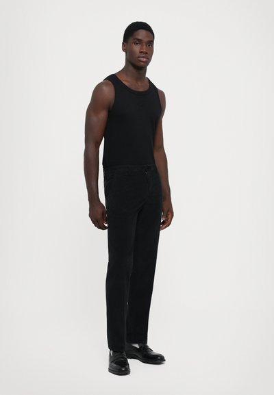 Joven vestido con una camiseta sin mangas negra, pantalones negros y zapatos de cuero negros, apoyado contra un fondo blanco liso.