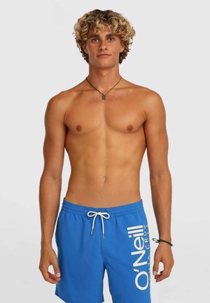 Jeune homme aux cheveux blonds bouclés portant un short de bain bleu O'Neill, un collier noir et un bracelet noir, debout devant un fond blanc.