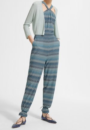 Frau trägt einen blau gemusterten Jumpsuit mit Zickzackmuster, eine leichte, glitzernde Kurzjacke und blaue Ballettschuhe mit Knöchelband.