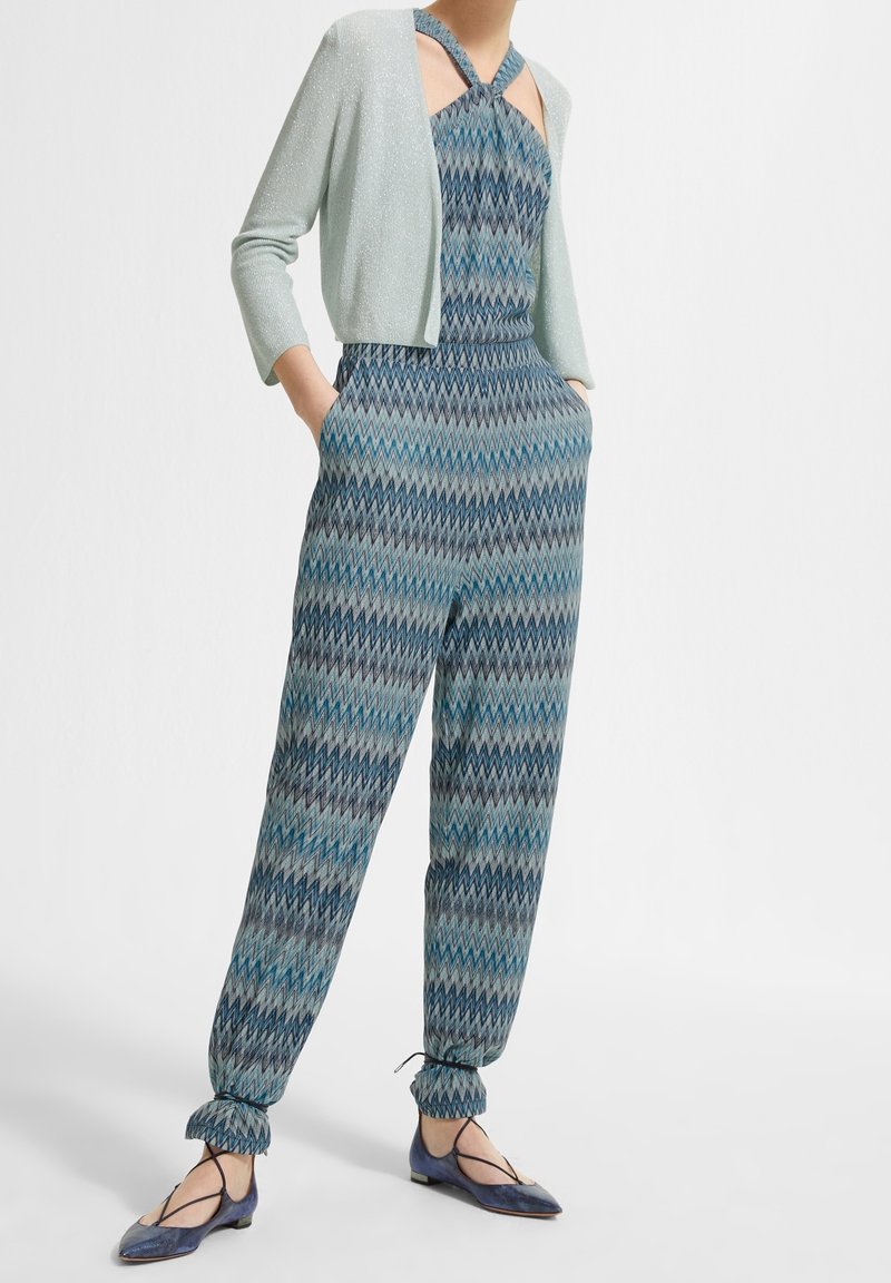 Femme portant une combinaison-pantalon bleue à motif zigzag, un cardigan court scintillant clair, et des ballerines bleues à liens chevilles.
