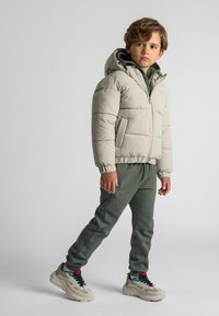Doudoune beige clair avec capuche, poignets élastiques et poches avant, associée à un pantalon de survêtement vert foncé et des baskets beiges volumineuses.