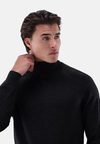 Mann, der den Kragen eines schwarzen Rollkragenpullis zurechtrückt, während er seitlich gegen einen schlichten weißen Hintergrund schaut.