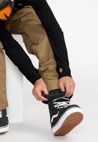 Bottes de neige noires avec des accents blancs, des lacets et des semelles en caoutchouc texturé. La personne ajuste le pantalon imperméable marron au niveau du poignet.