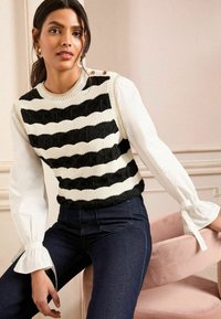Knitted black and white striped sweater with textured pattern, paired with a white shirt featuring flared sleeves and a pink chair.