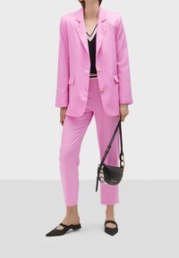 Rosa skräddarsydd kavaj och matchande byxor i ett mjukt tyg, med en svart v-ringad topp, svarta platta skor och en liten svart väska.