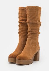 Bottes montantes en daim marron avec un design ample, bout rond et talon épais. Elles présentent une texture lisse et des détails de couture minimalistes.
