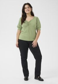 Green jersey top with puff sleeves, scooped neckline, and gathered detailing at the front, paired with black straight-leg pants and black shoes.