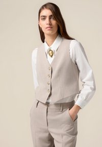 Beige maßgeschneiderte Weste mit fünf Knöpfen, getragen über einem weißen Hemd. Auffällige goldene Brosche am Kragen. Glattes Material und strukturierte Passform.