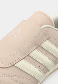 Gaiši rozā Adidas kedas ar trim krēmkrāsas svītrām, perforācijām un ievītām detaļām uz gludas ādas virsmas.