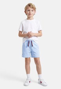 Νεαρός αγόρι που στέκεται με τα χέρια ενωμένα, φορώντας λευκό μπλουζάκι, ανοιχτόχρωμα μπλε σορτς με ριγέ κορδόνια, λευκές κάλτσες και λευκά αθλητικά παπούτσια.