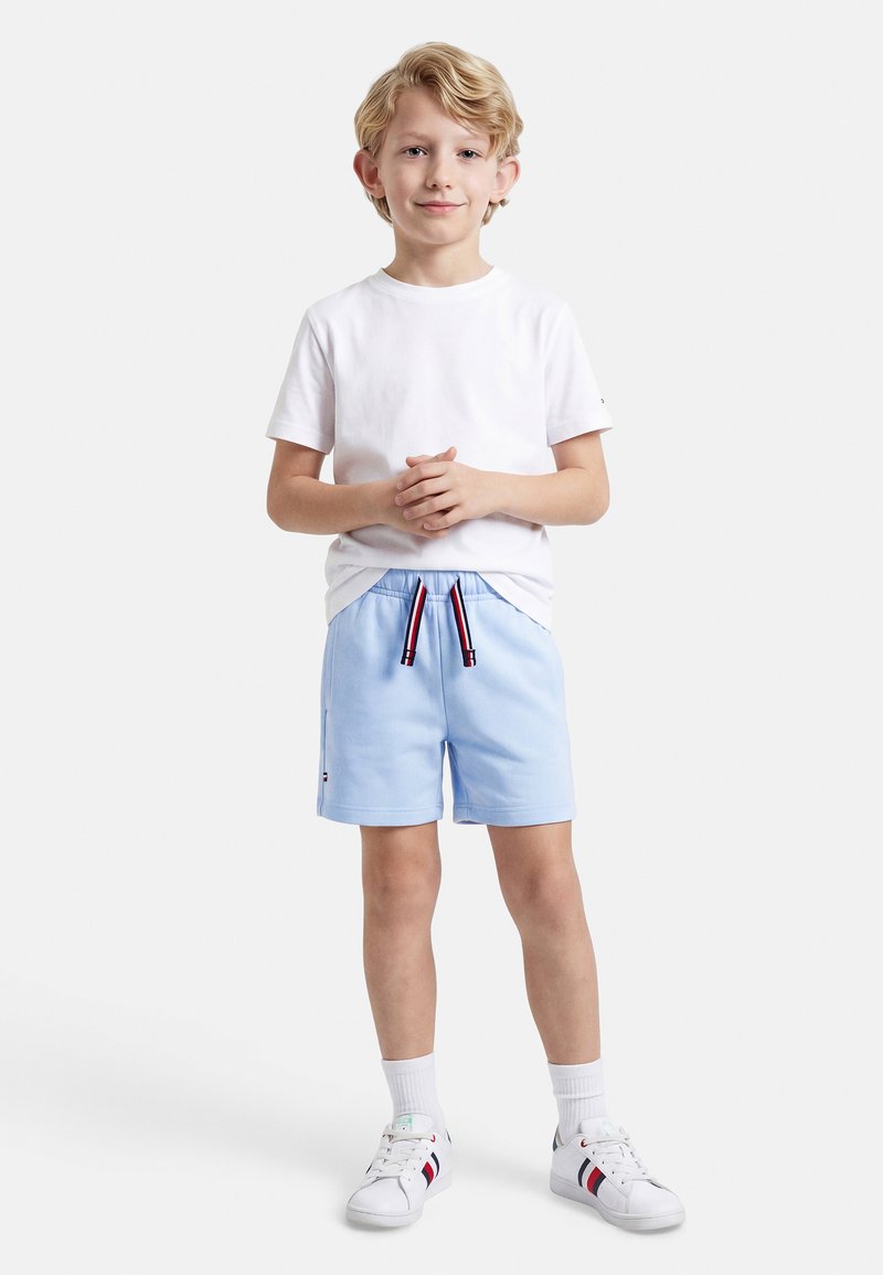 Νεαρός αγόρι που στέκεται με τα χέρια ενωμένα, φορώντας λευκό μπλουζάκι, ανοιχτόχρωμα μπλε σορτς με ριγέ κορδόνια, λευκές κάλτσες και λευκά αθλητικά παπούτσια.