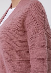 Ροζ πλεκτή ζακέτα με υφή ρίγες, που διαθέτει απαλό, χνουδωτό υλικό και ανοιχτό μπροστινό σχέδιο. Ορατά ράμματα και μοτίβα στα μανίκια.