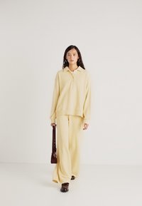 Ανοιχτό κίτρινο πλεκτό πουλόβερ με γιακά, συνδυασμένο με αντίστοιχες φαρδιές παντελόνες. Καφέ τσάντα κρατημένη στο δεξί χέρι, με λεία υφή σε όλη την επιφάνεια.