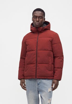 Mand iført en rød hættejakke med forlommer og lyseblå revnede jeans mod en ensfarvet hvid baggrund.