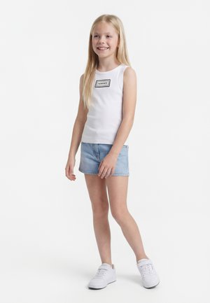 Jong meisje lacht, draagt een witte mouwloze top met "Versace"-logo, lichtblauwe spijkershorts en witte sneakers, staand op een witte achtergrond.