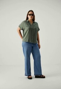 Donna che indossa occhiali da sole scuri, camicia verde con bottoni, jeans blu a gamba larga e sandali neri con zeppa, in piedi con una mano in tasca.