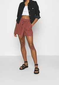 Brune højtaljede shorts med en bundet bælte og knapdetaljer, parret med en cropped hvid tanktop og sorte sandaletter med stropper. Sort denimjakke.