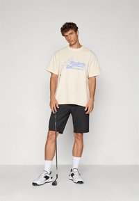 Smėlio spalvos medvilniniai marškinėliai su mėlynu užrašu, derinami su juodais šortais. Modelis laiko golfo lazdą, dėvi baltus ir juodus sportinius batelius.