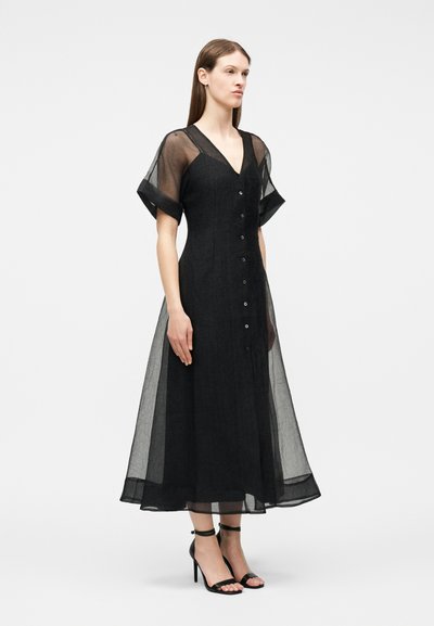 Γυναίκα στέκεται φορώντας μαύρο, κουμπωτό, διάφανο φόρεμα με κοντά μανίκια και V-λαιμόκοψη, μαζί με μαύρα σανδάλια με ψηλά τακούνια, πάνω σε απλό φόντο.