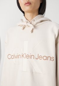 Felpa beige con tasca a canguro, cappuccio con cordino e logo in rilievo "Calvin Klein Jeans" in contrasto beige. Morbido al tatto.