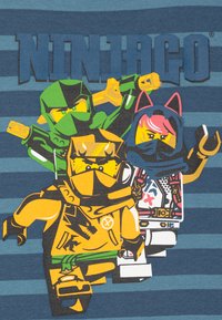 Lego Ninjago grafisk T-skjorte med blå og grå stripe bakgrunn. Har tre fargerike ninja-figurer i detalj med tilbehør og mønstre.