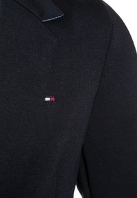 Černá pletená sako se strukturovaným límcem. Na levé straně hrudi se nachází malá červená, bílá a modrá logo, látka má texturovaný povrch.