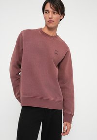 Sudadera burdeos con puños y dobladillo acanalados, tela texturizada y un sutil logo impreso en el pecho, combinada con pantalones negros.