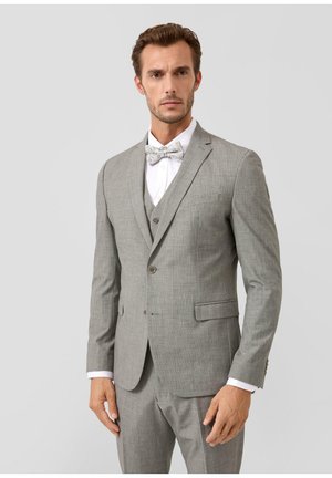 Homme portant un costume trois pièces gris clair avec une chemise blanche et un nœud papillon à motifs, debout devant un fond uni gris clair.