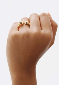 Anillos de color dorado con esferas pulidas de diferentes tamaños, llevados en una mano con una textura de piel suave sobre un fondo claro.