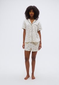 Σετ πιτζάμας σε ανοιχτό μπεζ χρώμα με κοντά μανίκια και σορτς, διακοσμημένο με μικρό μαύρο σχέδιο και αντίθετη μαύρη επένδυση στα άκρα.