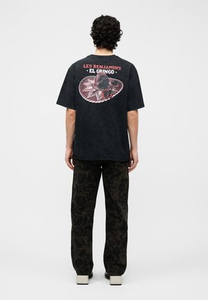 Άνδρας που στέκεται με την πλάτη στην κάμερα, φοράει μαύρο T-shirt με κόκκινη και λευκή επιγραφή "LES BENJAMINS EL GRINGO" και γραφικό καουμπόικου καπέλο, σκούρο παντελόνι με σχέδια, λευκά παπούτσια.