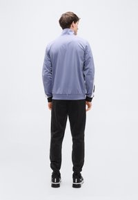 Αθλητικό τζάκετ ανοιχτού μπλε με ψηλό γιακά, λωρίδες στο πλάι και ριμπ μανσέτες, σε συνδυασμό με μαύρο παντελόνι και μαύρα αθλητικά παπούτσια.